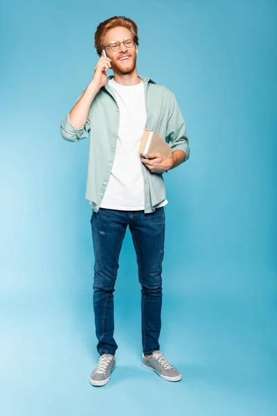 Pelirroja y barbuda estudiante en gafas hablando en smartphone y sosteniendo libros en azul - foto de stock