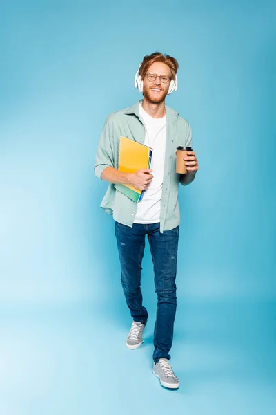 Estudiante barbudo en gafas y auriculares inalámbricos sosteniendo taza de papel y cuadernos mientras camina sobre azul - foto de stock