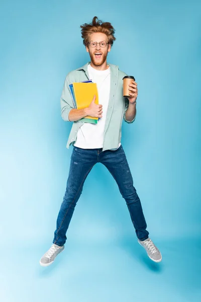 Estudiante barbudo en gafas sosteniendo taza de papel y cuadernos mientras salta sobre azul - foto de stock