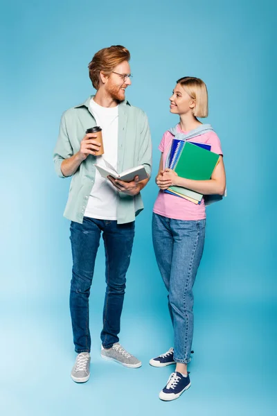 Rothaarige Studentin hält Buch und Coffee to go in der Hand, während sie eine blonde junge Frau mit Notizbüchern auf blauem Grund ansieht — Stockfoto