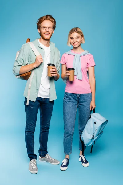 Jóvenes estudiantes sosteniendo café para ir y mochilas mientras están de pie en azul - foto de stock