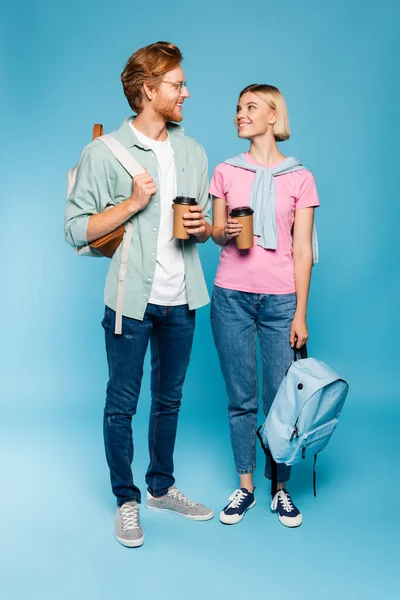 Jóvenes estudiantes sosteniendo vasos de papel y mochilas mientras están de pie en azul - foto de stock