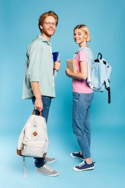 Young students with backpacks holding notebooks and books while standing on blue — Stock Photo