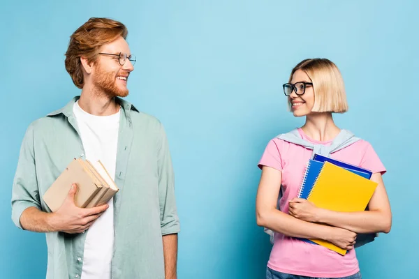 Giovani studenti in occhiali che tengono quaderni e libri mentre si guardano l'un l'altro su blu — Foto stock