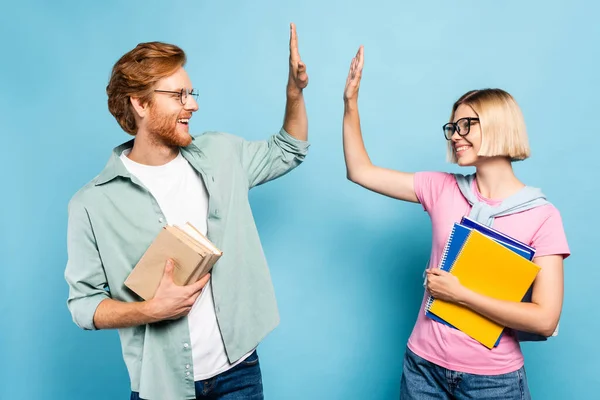 Young students in glasses holding notebooks and books while giving high five on blue — Stock Photo