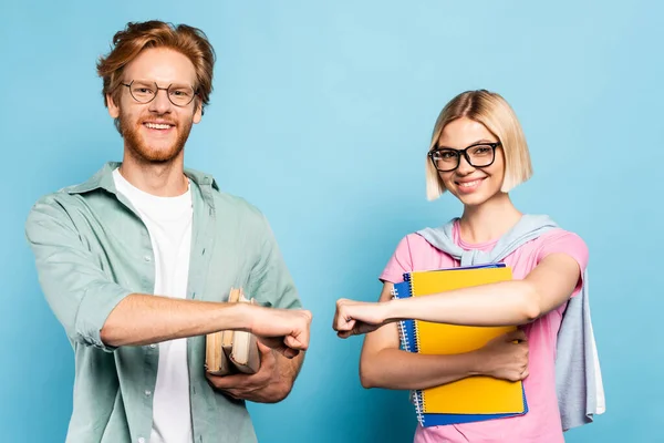Young students in glasses holding notebooks and books while fist bumping on blue — Stock Photo