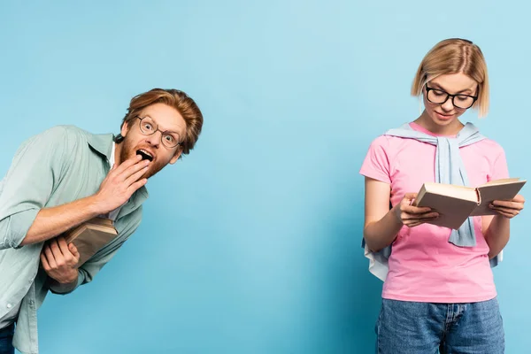 Surprised and redhead man looking at camera while blonde woman reading book on blue — Stock Photo