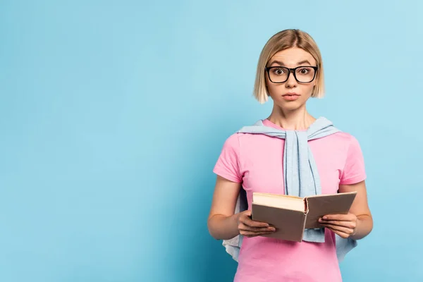 Estudiante rubia en gafas sosteniendo libro y mirando a la cámara en azul - foto de stock