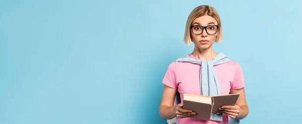 Panoramic concept of blonde student in glasses holding book and looking away on blue — Stock Photo