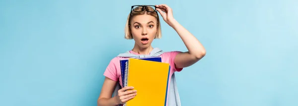 Panoramaaufnahme eines schockierten Studenten, der eine Brille berührt, während er Notizbücher auf blau hält — Stockfoto