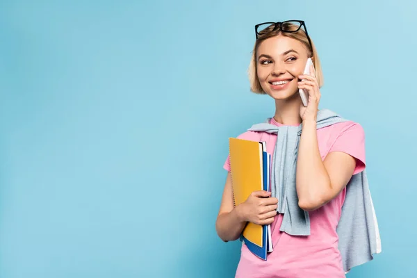 Joven estudiante rubia sosteniendo cuadernos y hablando en el teléfono inteligente en azul - foto de stock