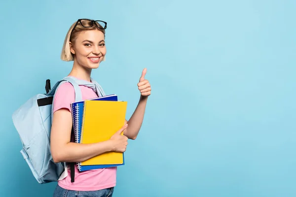 Blonde Studentin mit Rucksack hält Notizbücher in der Hand und zeigt Daumen hoch auf blau — Stockfoto