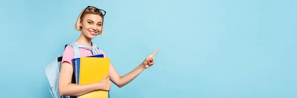 Panoramic crop of young blonde student with backpack holding notebooks and pointing with finger on blue — Stock Photo
