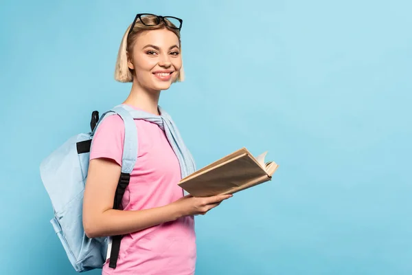 Junge blonde Studentin mit Rucksack hält Buch auf blau mit Kopierraum — Stockfoto