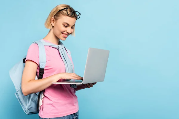 Young blonde student with backpack using laptop on blue — Stock Photo