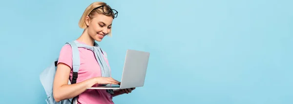 Panoramic concept of young blonde student with backpack using laptop isolated on blue — Stock Photo