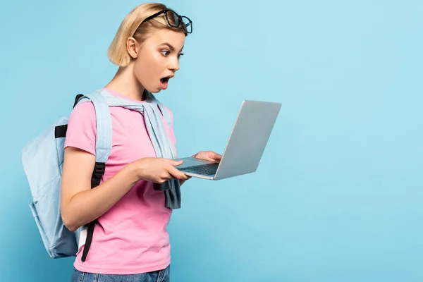 Shocked blonde student with backpack using laptop on blue — Stock Photo