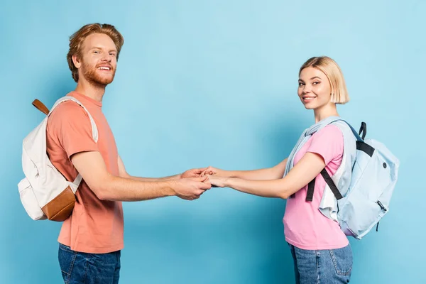 Redhead and blonde students with backpacks looking at camera and holding hands on blue — Stock Photo
