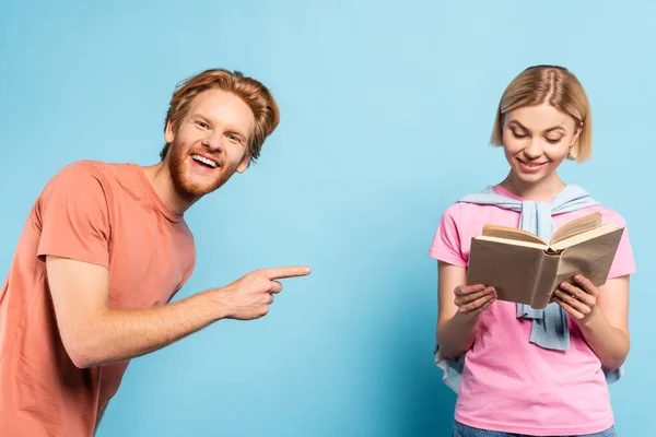 Bärtige Studentin zeigt mit dem Finger auf blonde Frau, die Buch auf blauem Grund liest — Stockfoto