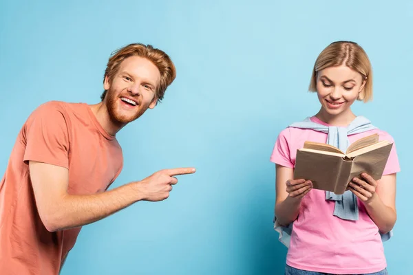 Estudiante barbudo señalando con el dedo a la mujer rubia leyendo libro sobre azul - foto de stock