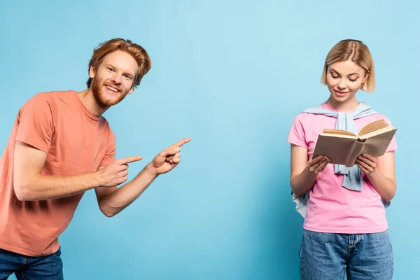 Bearded student pointing with fingers at blonde woman reading book on blue — Stock Photo