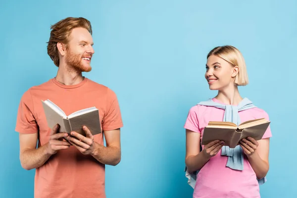Redhead and blonde students looking at each other while holding books on blue — Stock Photo