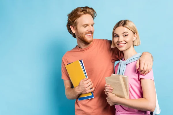 Redhead man holding notebooks and hugging blonde student with books on blue — Stock Photo