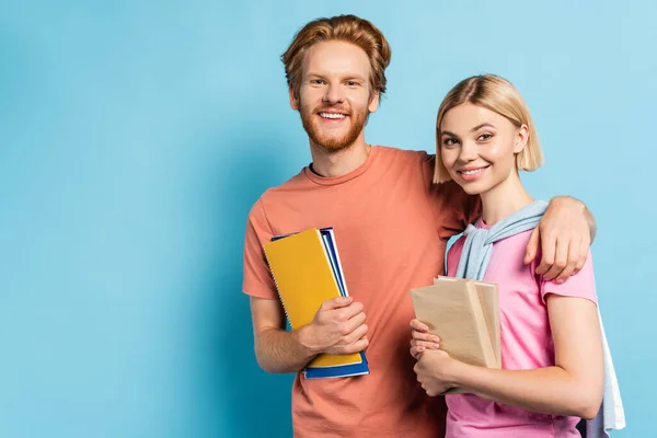 Bearded man holding notebooks and hugging blonde student with books on blue — Stock Photo