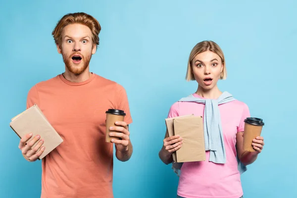 Shocked young students holding books and paper cups on blue — Stock Photo