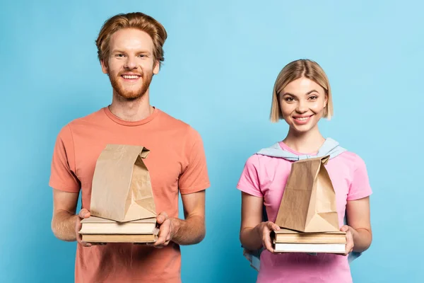 Redhead and blonde students holding paper bags and books on blue — Stock Photo