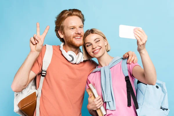 Enfoque selectivo de estudiante pelirroja con mochila mostrando signo de paz mientras toma selfie con amigo en azul - foto de stock