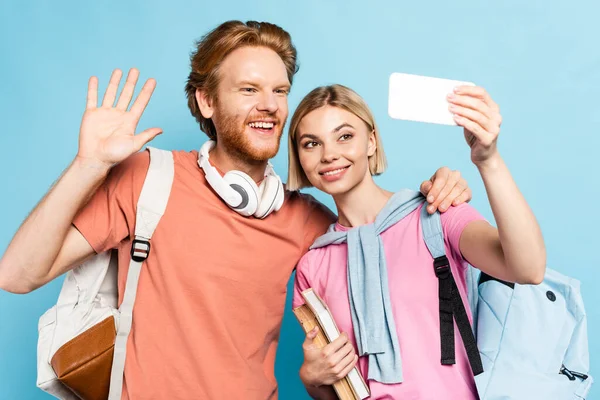 Selective focus of redhead student with backpack waving hand while taking selfie with friend on blue — Stock Photo