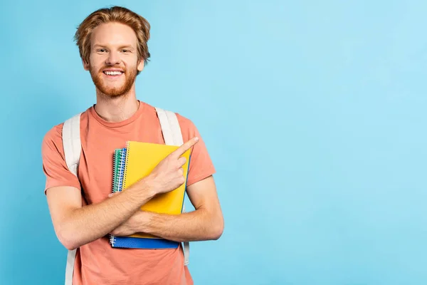 Pelirroja estudiante sosteniendo cuadernos y señalando con el dedo en azul - foto de stock