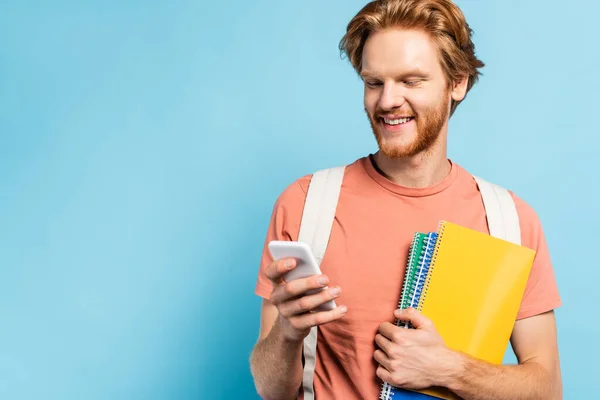 Rousse étudiant tenant des cahiers et utilisant smartphone sur bleu — Photo de stock