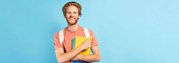 Panoramic shot of redhead student holding notebooks and pointing with finger on blue — Stock Photo