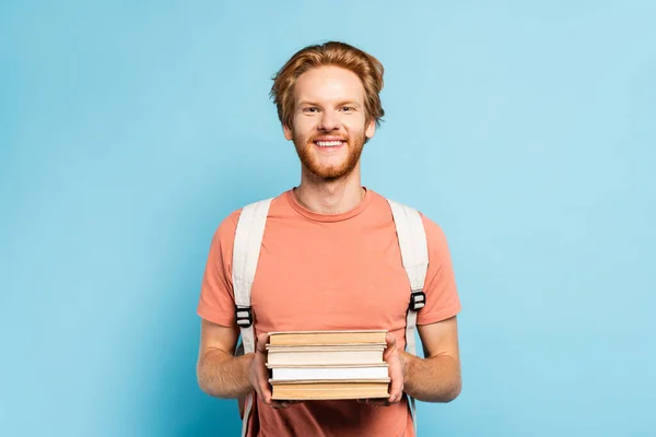 Pelirroja estudiante sosteniendo libros y mirando a la cámara en azul - foto de stock