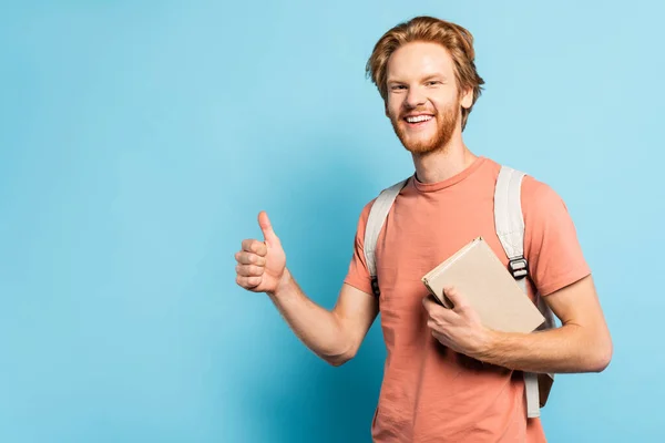 Bearded student holding book and showing thumb up on blue — Stock Photo