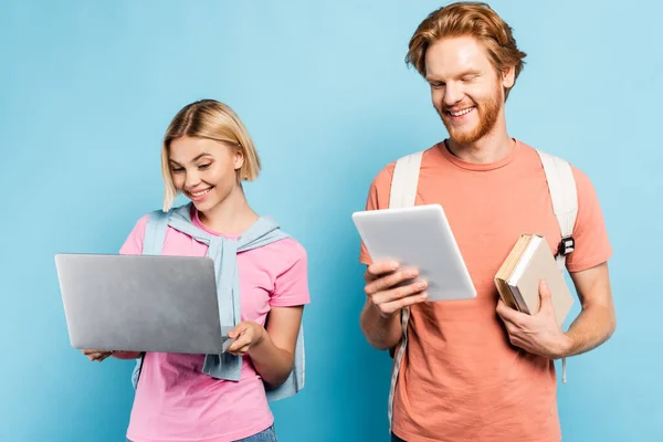 Estudiante pelirroja sosteniendo libros y usando tableta digital cerca de amigo rubio con portátil en azul - foto de stock