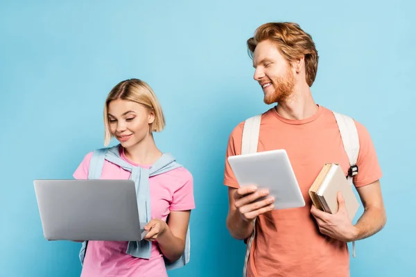 Estudiante pelirroja sosteniendo libros y tableta digital mientras mira a un amigo rubio usando el portátil en azul - foto de stock