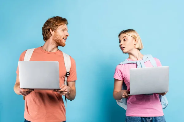 Curieux jeunes étudiants regardant les uns les autres ordinateurs portables sur bleu — Photo de stock