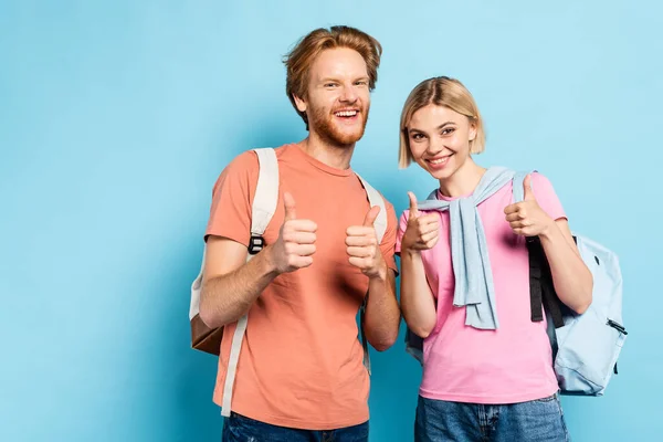 Jóvenes estudiantes con mochilas mostrando pulgares hacia arriba en azul - foto de stock
