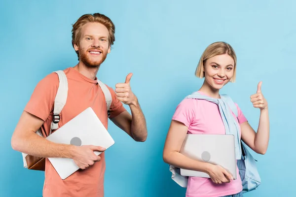 Jóvenes estudiantes sosteniendo computadoras portátiles y mostrando los pulgares hacia arriba en azul - foto de stock