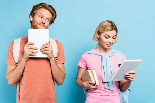 Pelirrojo hombre mirando rubia estudiante sosteniendo libros y tableta digital en azul - foto de stock