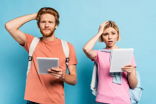 Pensive students holding digital tablets and touching heads on blue — Stock Photo