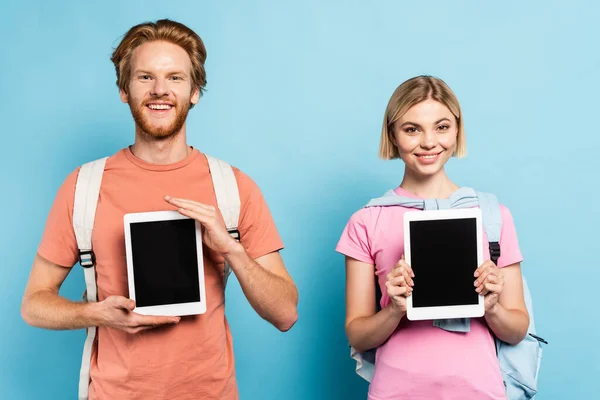 Blonde und rothaarige Studenten halten digitale Tablets mit leerem Bildschirm auf blauem Hintergrund — Stockfoto