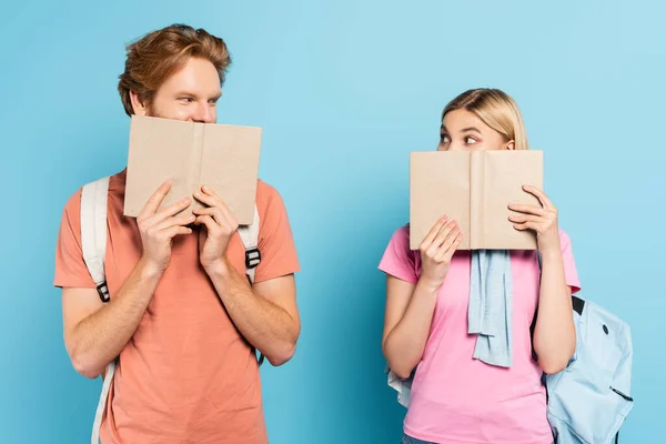 Jovens estudantes olhando uns para os outros enquanto cobria rostos com livros sobre azul — Fotografia de Stock