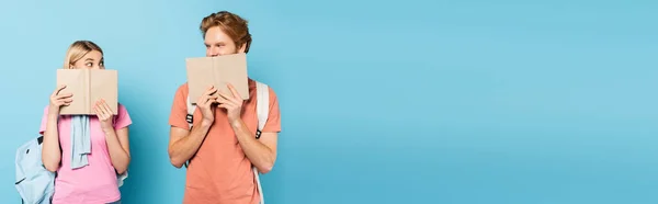 Horizontal image of young students looking at each other while covering faces with books on blue — Stock Photo