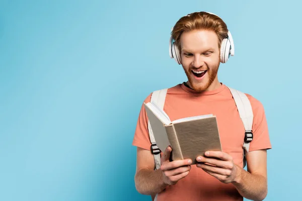 Studente eccitato in cuffie wireless lettura libro sul blu — Foto stock