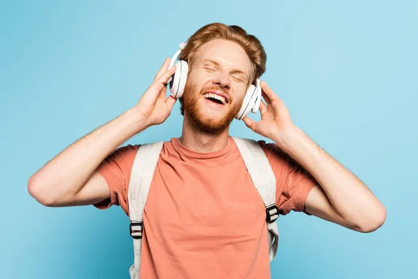 Étudiant rousse avec les yeux fermés touchant écouteurs sans fil sur bleu — Photo de stock