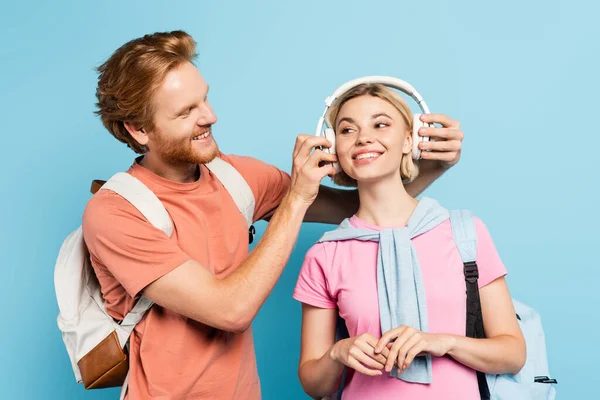 Bärtiger Student mit Rucksack, der drahtlose Kopfhörer einer blonden Freundin auf blauem Grund berührt — Stockfoto